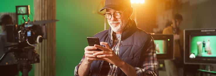 Individual who could be a director or producer, looking at their mobile phone while on set with cameras and green screens in the background.